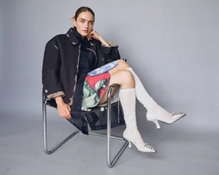 Model wearing a black jacket and white heels seated on a chrome chair against a grey seamless background
