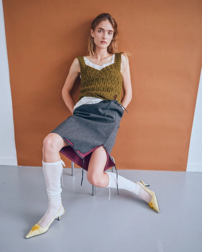 Model in olive knit tank top, grey skirt, white knee socks and yellow heels seated against a terracotta studio backdrop.
