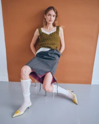 Model in olive knit tank top, grey skirt, white knee socks and yellow heels seated against a terracotta studio backdrop.