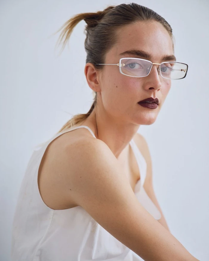 Model wearing thin-framed rectangular glasses, white top, and dark lipstick against a plain white background