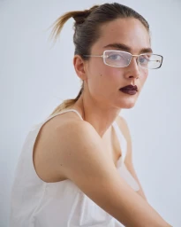 Model wearing thin-framed rectangular glasses, white top, and dark lipstick against a plain white background