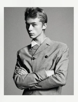 Monochrome portrait of a young male model with folded arms wearing a grey blazer and white shirt against a plain grey backdrop.