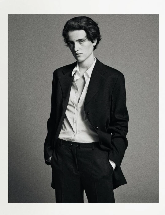 Model wearing a dark tailored suit jacket and matching trousers with a white buttoned shirt against a textured grey background.
