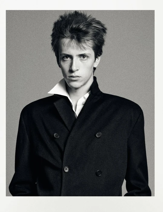 Portrait of a young man wearing a black double-breasted coat and a white collared shirt against a grey studio backdrop.