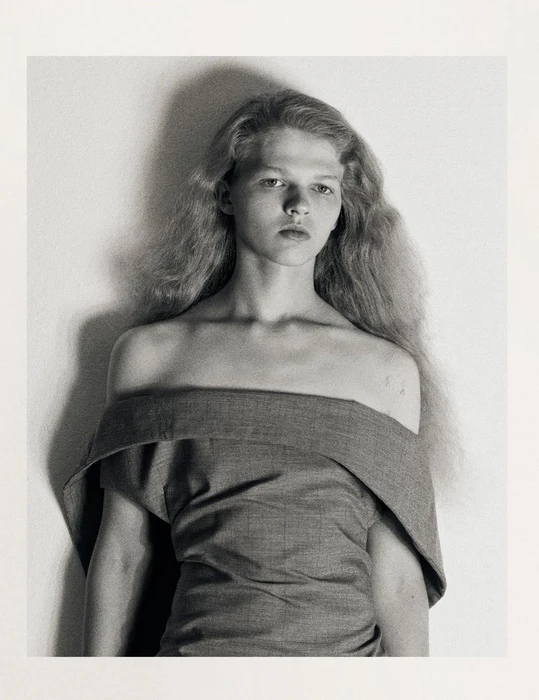 Portrait of a model with long hair wearing an off-shoulder grey top against a textured plain wall.