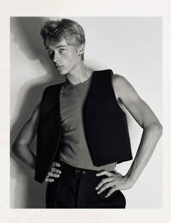 Young male model posing in a black vest and dark trousers against a white seamless studio backdrop in a black and white portrait