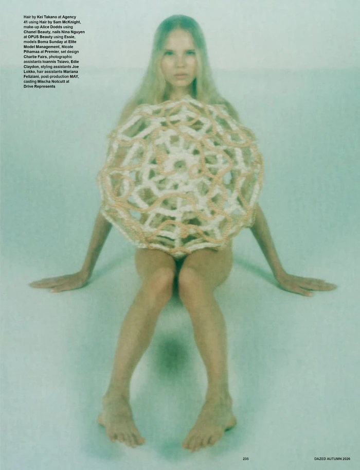 Model seated on a pale surface holding an intricate woven textile piece in front of her torso against a soft teal background