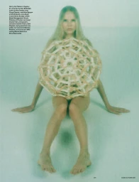 Model seated on a pale surface holding an intricate woven textile piece in front of her torso against a soft teal background