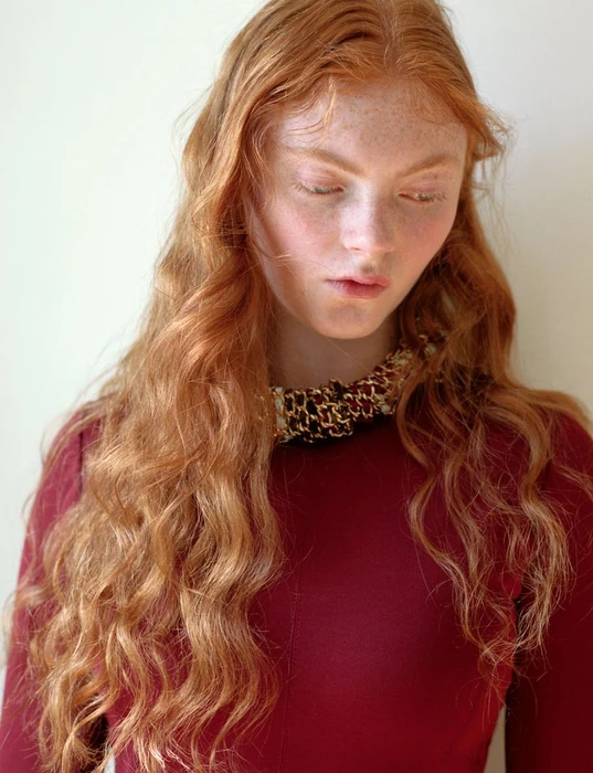 Red-haired female model with freckles wearing a burgundy top and ornate beaded necklace against a plain white background