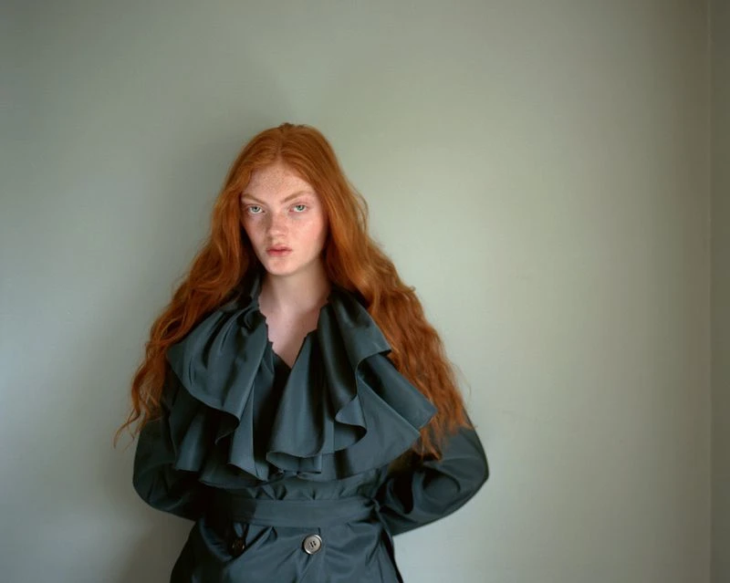 Red-haired female model wearing a dark ruffled coat against a plain pale wall