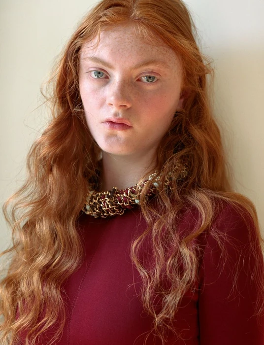 Close-up portrait of a red-haired woman wearing a textured burgundy necklace against a plain neutral background.