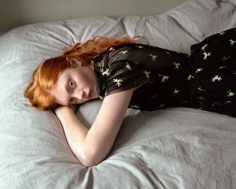 Red-haired model lying on a grey bed wearing a dark sheer dress with embroidered star patterns