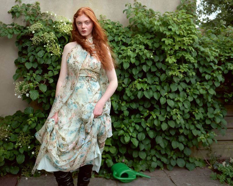 Red-haired model in a floral halterneck dress and black boots standing outdoors in front of a leafy green bush.