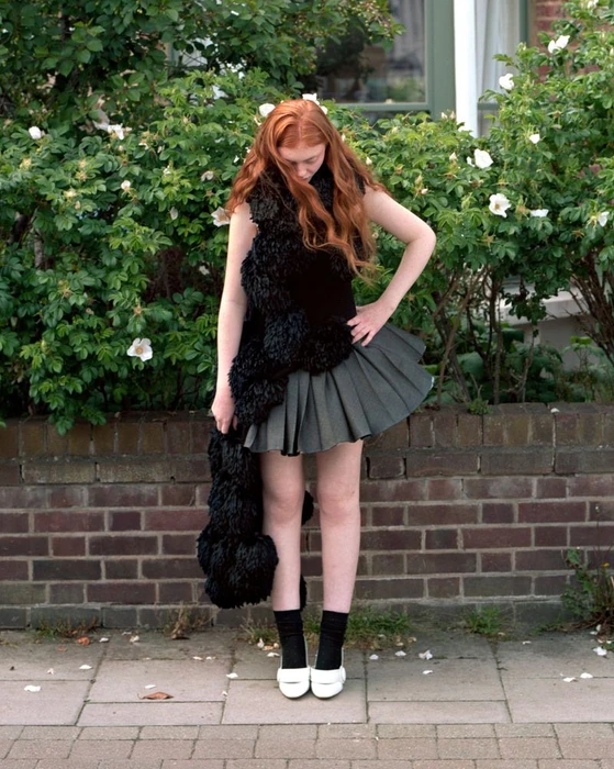 Red-haired model wearing a black textured wrap and pleated grey skirt in front of flowering bushes.