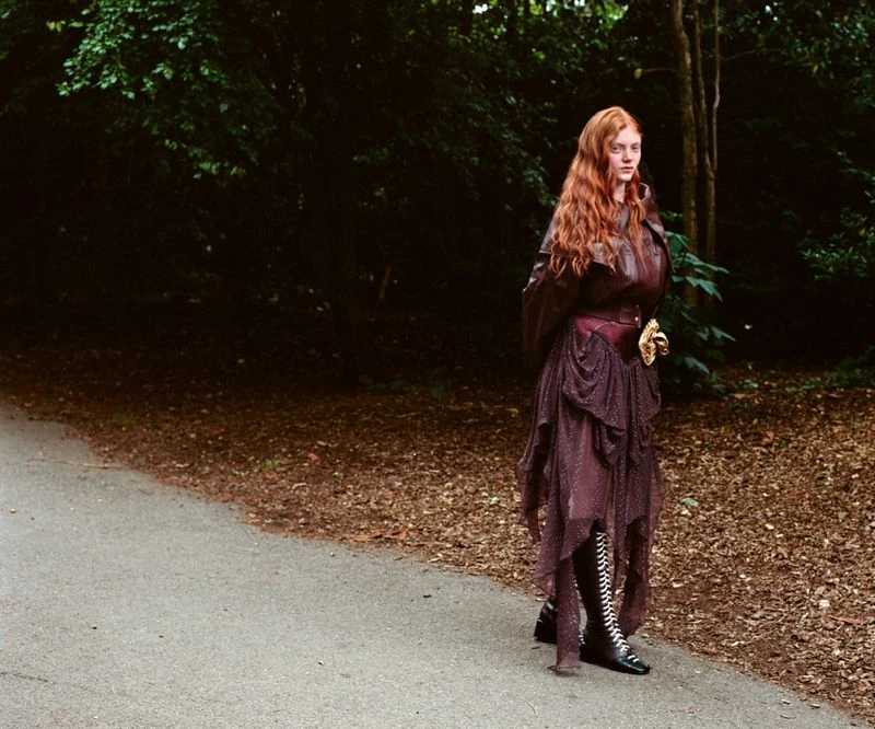 Red-haired female model standing on an asphalt path wearing a dark leather jacket and layered burgundy dress