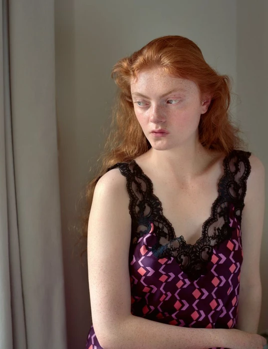 Red-haired female model wearing a patterned purple camisole with black lace trim standing near a window.