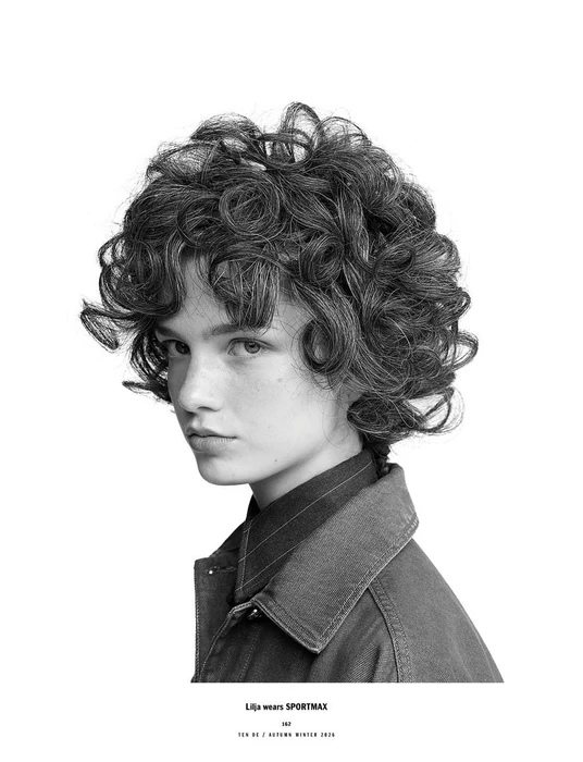Monochrome portrait of model Lilja Drab with curly hair wearing a dark collared jacket against a white background for 10 Magazine