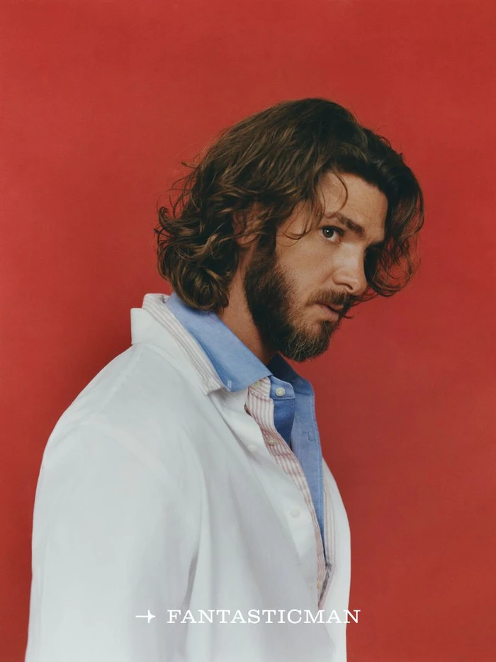 Actor Andrew Garfield with long curly hair and beard wearing a white coat and blue shirt against a solid red background