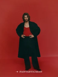 Andrew Garfield posing against a solid red backdrop wearing a red tank top and black coat