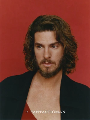 Portrait of Andrew Garfield with medium-length wavy brown hair and beard against a solid red backdrop