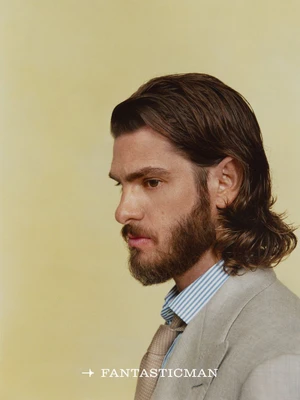 Profile portrait of Andrew Garfield with medium-length dark hair and a beard, wearing a grey suit jacket, blue-striped shirt, and beige tie against a yellow background.