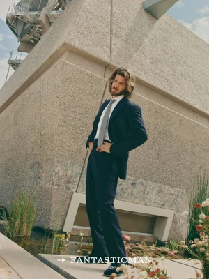 Andrew Garfield posing outdoors in a dark suit and grey tie in front of a modern concrete building structure