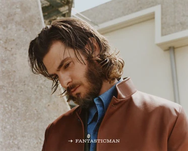 Andrew Garfield with long hair and beard wearing a brown leather jacket and blue shirt against a building exterior.