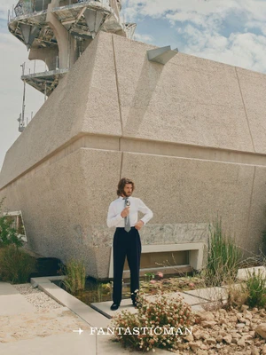 Andrew Garfield posing in a white shirt and dark trousers outdoors against a brutalist architectural structure.