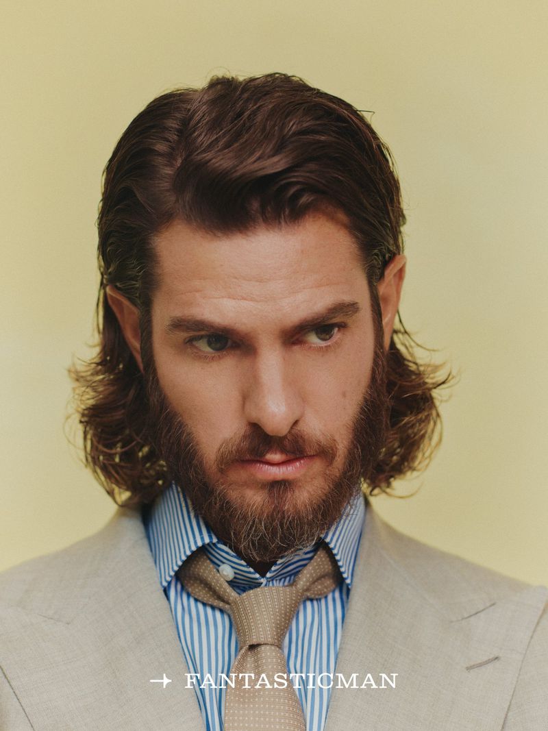 Portrait of Andrew Garfield with long hair and a beard wearing a striped shirt and tan tie against a pale background