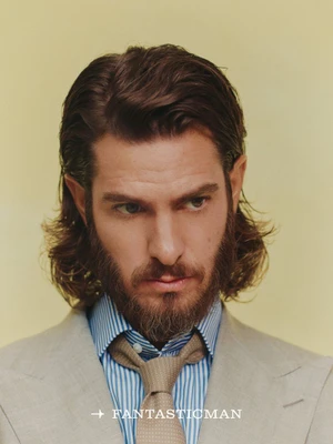 Portrait of Andrew Garfield with long hair and a beard wearing a striped shirt and tan tie against a pale background