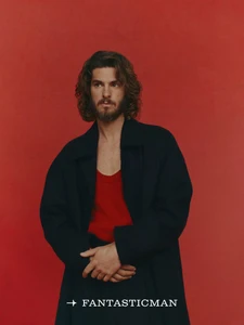 Andrew Garfield with long hair and beard wearing a dark coat and red top against a red background