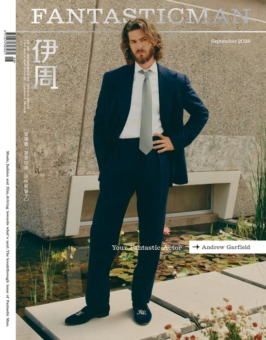 Andrew Garfield posing in a dark blue suit with a white shirt and grey tie in front of a concrete building and water garden.