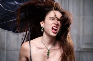 Model with long brown hair blowing in the wind holding an open umbrella while wearing a gemstone necklace and matching earrings