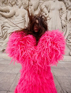 Model in vibrant hot pink feathered dress standing in front of ornate classical sculptural relief artwork