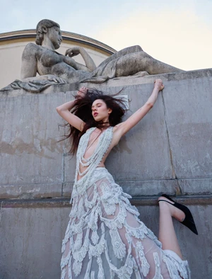 Model wearing a sheer embellished gown posing below a classical stone statue for ICON Magazin Germany.