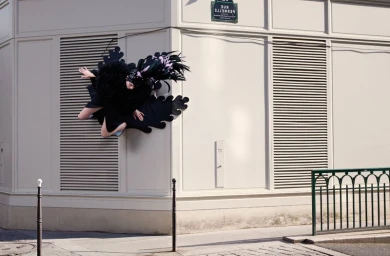 Model in black feathered outfit suspended against the corner of a light building exterior