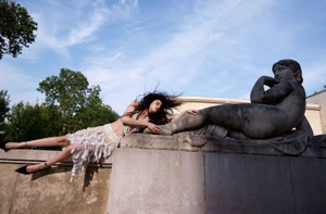 Model Isadora Ribeiro lying on a stone wall next to a large classical female statue under a clear sky for ICON Magazin Germany.