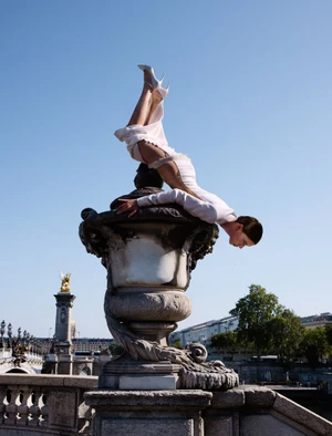 Model balanced horizontally on an ornate stone bridge urn against a clear blue sky for ICON Magazin Germany.