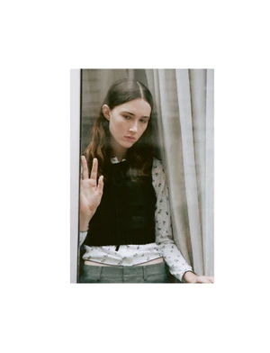 Young woman wearing a patterned white blouse and black vest posing indoors near sheer curtains