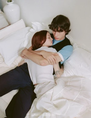 Young couple embracing in bed, woman with dark hair wearing white top, man wearing blue t-shirt and black vest.