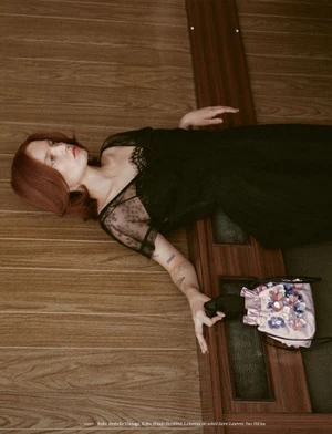 Woman in black lace dress lying on a wood paneled floor holding a pink embellished handbag