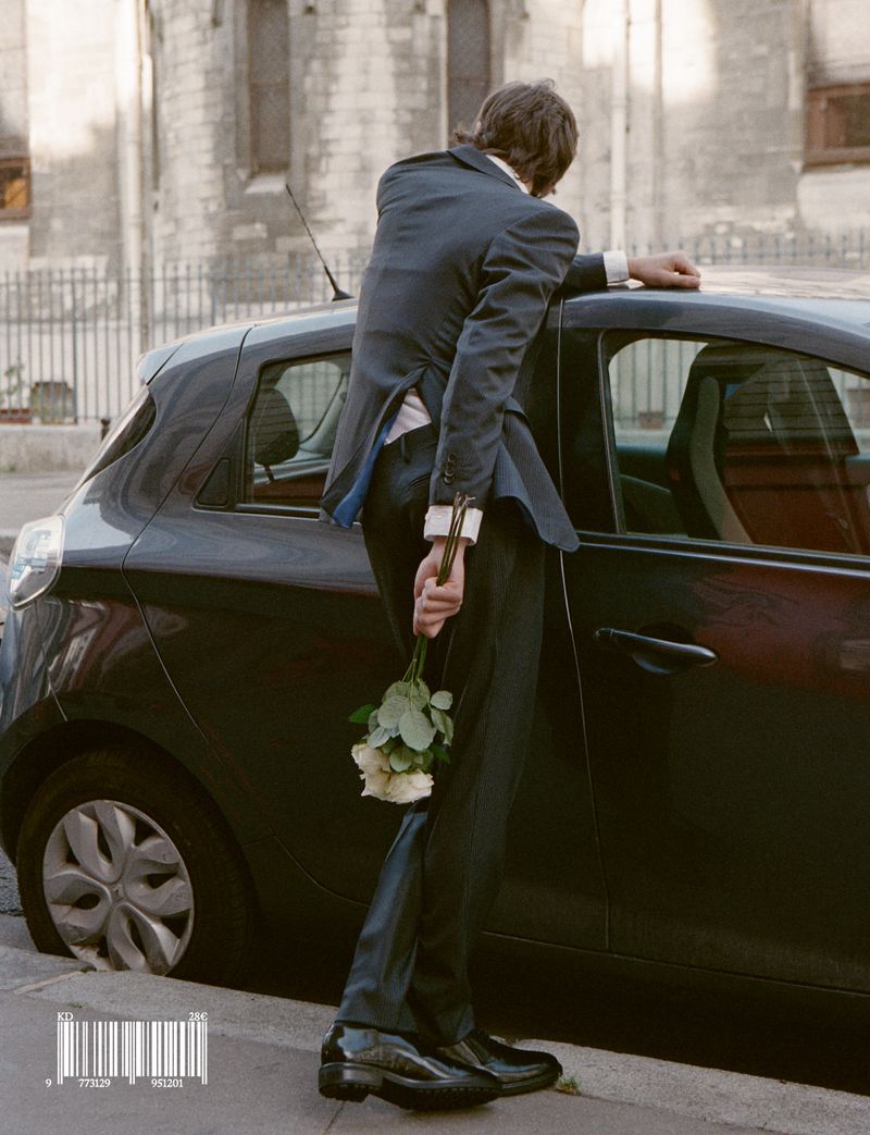 A man in a dark suit leaning against a car holding a bouquet of white roses behind his back on a street.