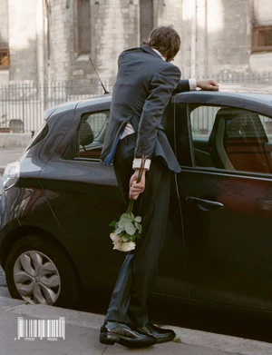 A man in a dark suit leaning against a car holding a bouquet of white roses behind his back on a street.