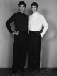 Two male models posing side by side against a neutral studio background, wearing contrasting black and white shirts with dark trousers.