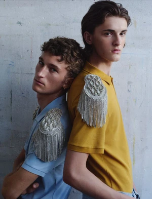 Two male models standing back-to-back wearing pastel polo shirts embellished with large silver crystal epaulettes.