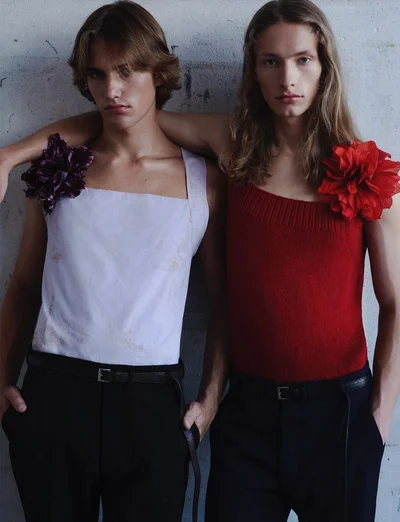 Two male models posing side by side wearing sleeveless tops adorned with large floral pins against a concrete background.