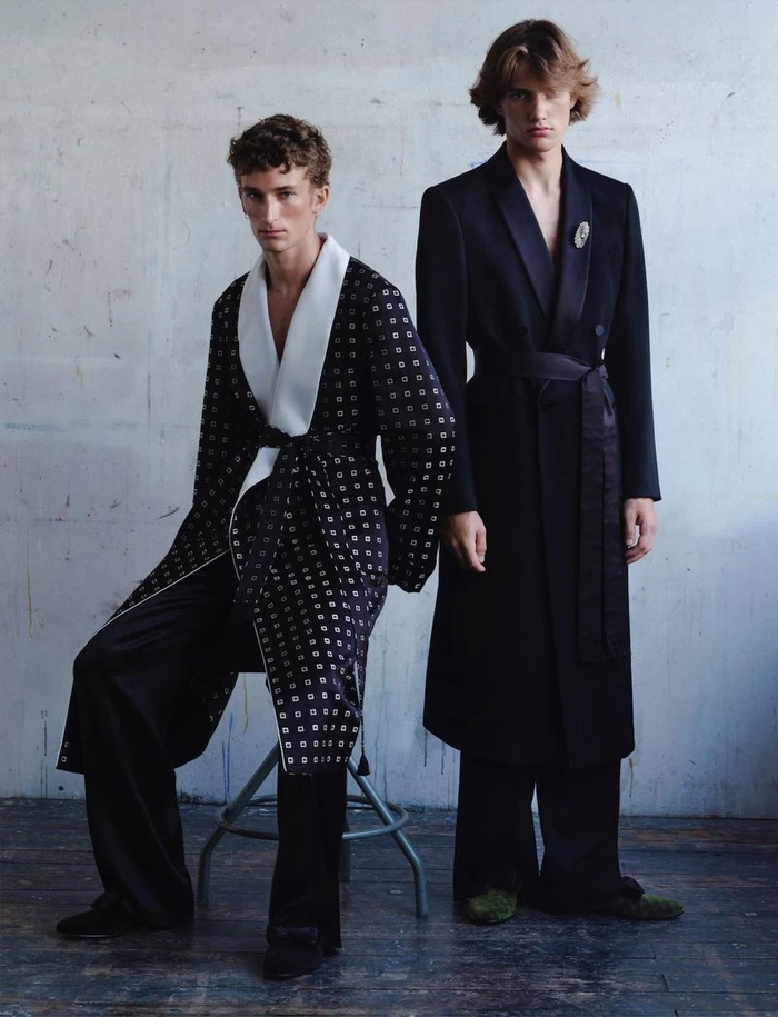 Two male models posing against a textured wall, one seated in a patterned robe and the other standing in a belted black coat.