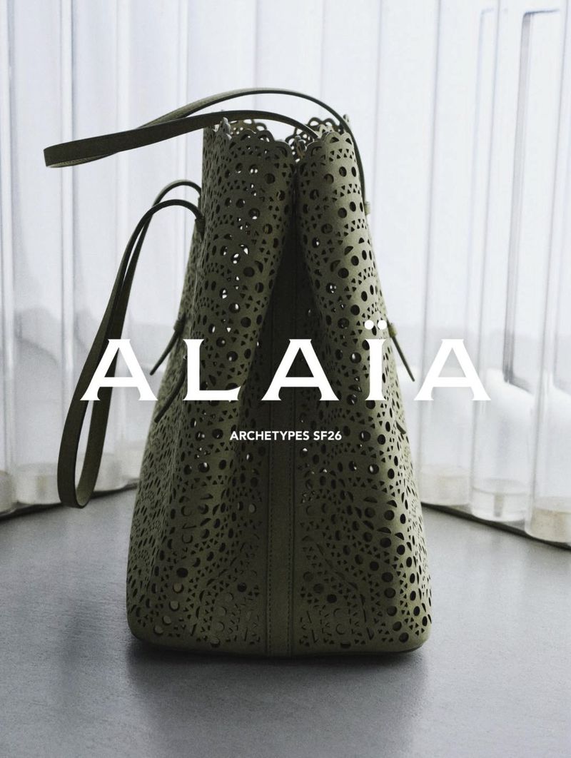 Close-up of an olive green perforated leather tote bag standing upright on a grey surface against a translucent white backdrop.