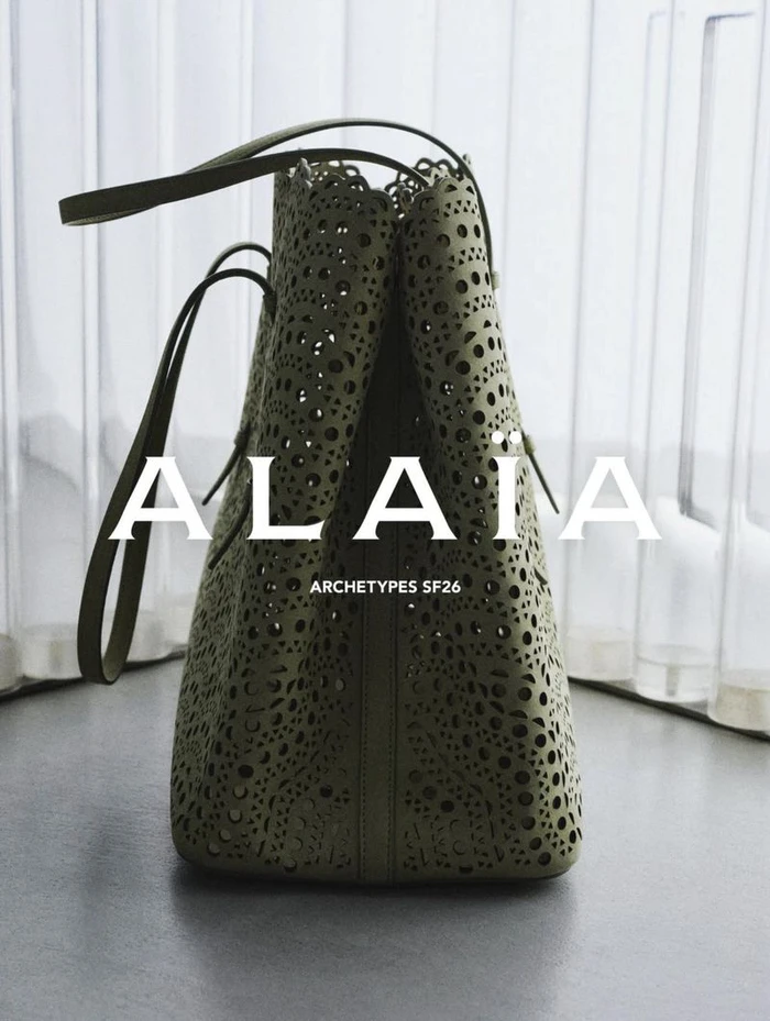 Close-up of an olive green perforated leather tote bag standing upright on a grey surface against a translucent white backdrop.
