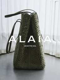 Close-up of an olive green perforated leather tote bag standing upright on a grey surface against a translucent white backdrop.
